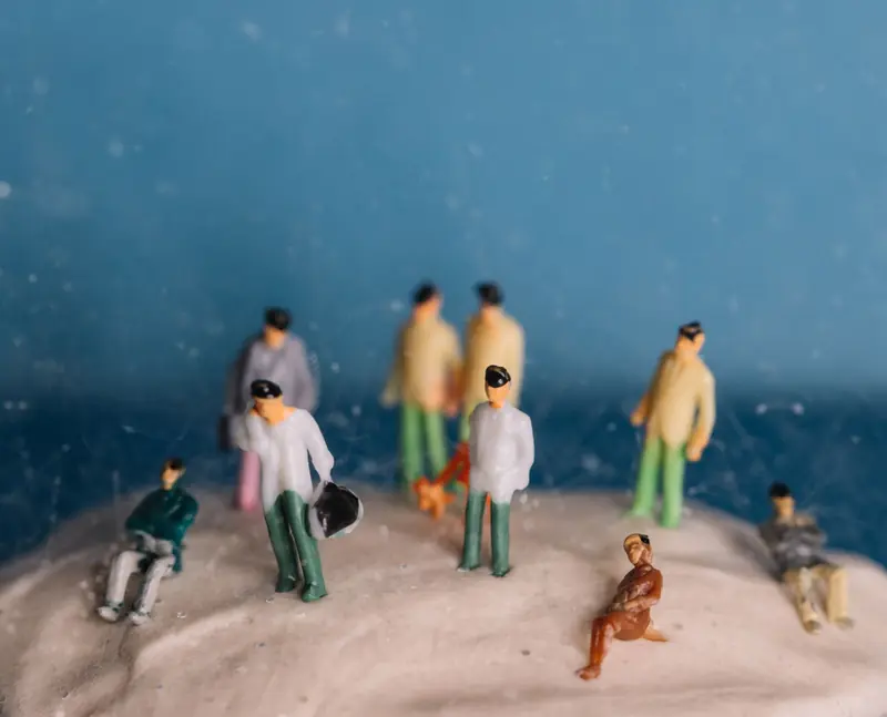 Miniature people figurines on textured surface with blue backdrop, maintaining a whimsical still life theme