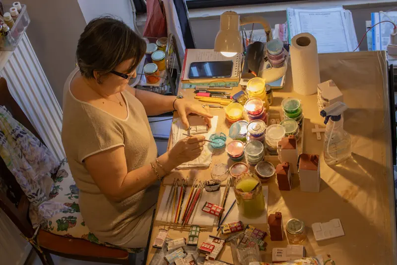 A woman meticulously painting miniature models at a cluttered art table in a well-lit studio